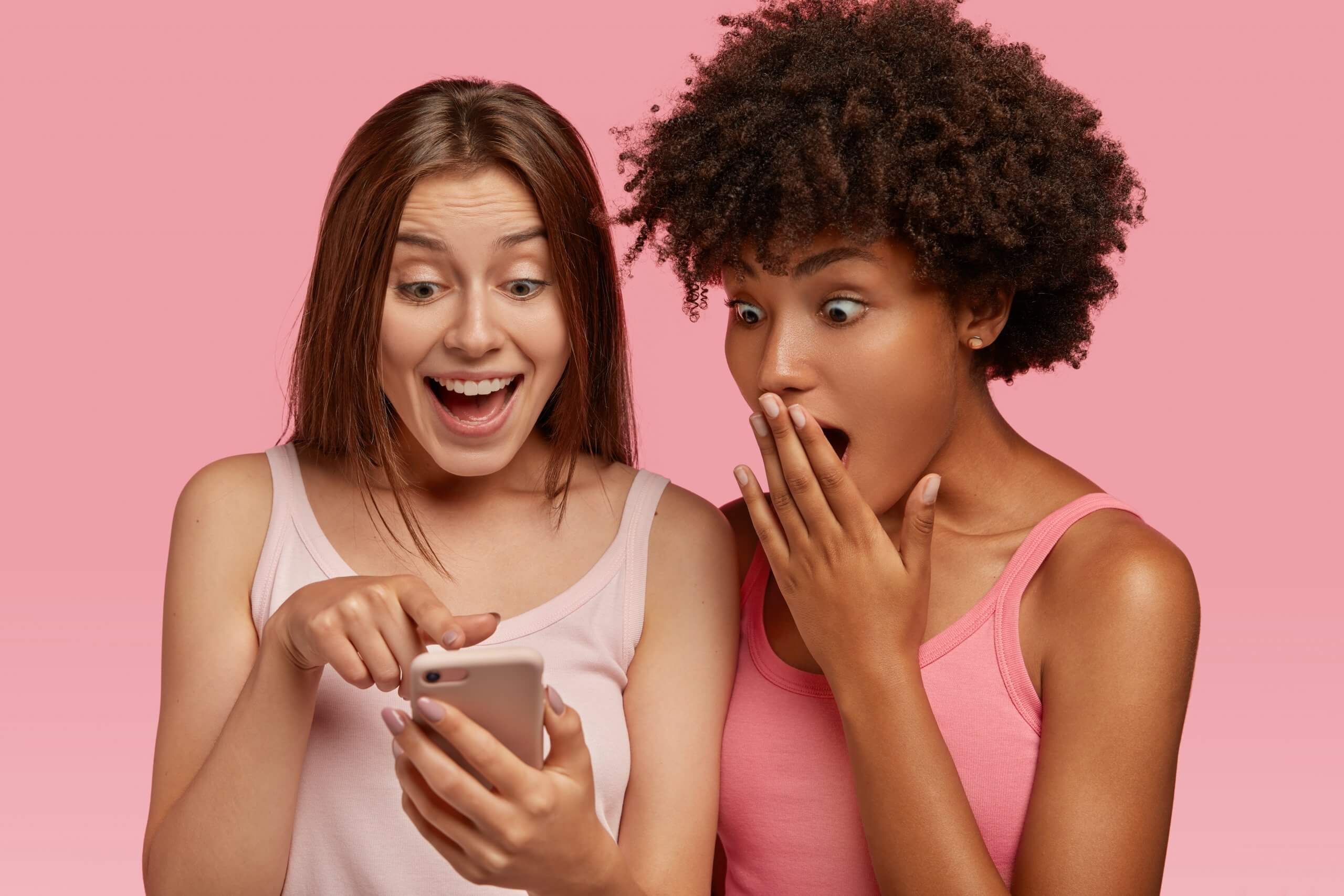 [:es]Two multiethnic young girls check out amazing article on web page via cellular, have surprised happy expressions, dressed in summer clothes, use wireless internet, model against pink background[:]